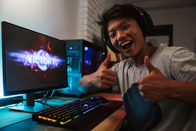 暗い部屋でコンピューターでビデオゲームをプレイし ヘッドフォンを着用し バックライト付きのカラフルなキーボードを使用しながら勝利を喜んでいる若いアジアの ゲーマー少年 プレミアム写真