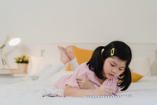 自宅で描く若いアジアの女の子 アジア日本人女性子供子供は ベッドの上に横たわる寝る前にスケッチブックで楽しい楽しい幸せ漫画をリラックス 夜のコンセプトで寝室で快適さと落ち着きを感じるリラックスします 無料の写真