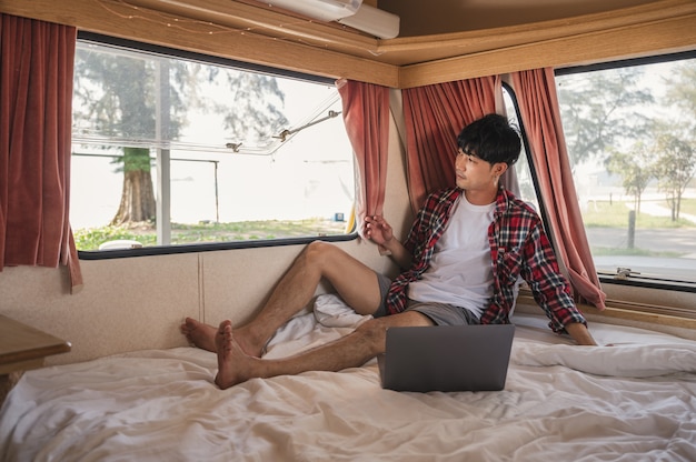 Young asian man wearing scott shirt relaxing with laptop on the bed in camper van Premium Photo