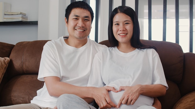Free Photo | Young asian pregnant couple making heart sign holding belly.  mom and dad feeling happy smiling peaceful while take care baby, pregnancy  lying on sofa in living room at home .