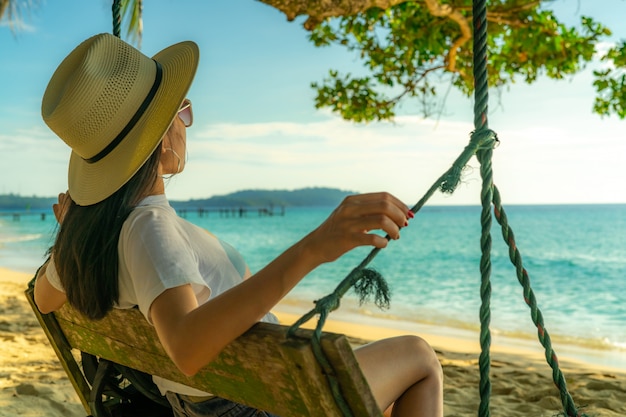 アジアの若い女性が座って 夏休みに海辺のブランコでリラックスします 夏のバイブ 女性は休日に一人で旅行します 熱帯の楽園のビーチでのバックパッカー プレミアム写真