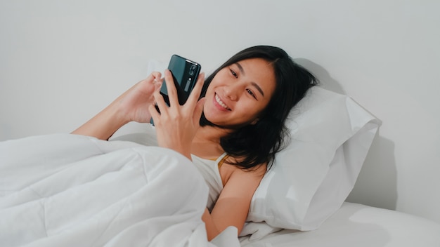 Young asian woman using smartphone checking social media feeling happy smiling while lying on bed after wake up in the morning, beautiful attractive hispanic lady smiling relax in bedroom at home. Free Photo