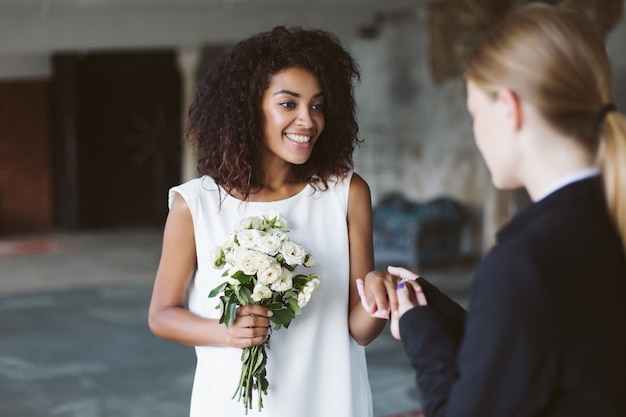 結婚式に楽しく時間を過ごしながら 花の小さな花束を手に持っている白いドレスの暗い巻き毛を持つ若い魅力的なアフリカ系アメリカ人の女性 プレミアム写真