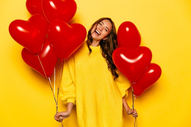 Young attractive girl with long curly hair, in yellow sweater holding red air balloons Free Photo