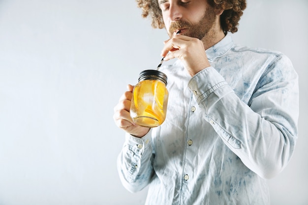 Young bearded man in light jeans shirt drinks fresh home made orange citrus sparkling lemonade through striped drinking straw from rustic transparent jar in hands Free Photo