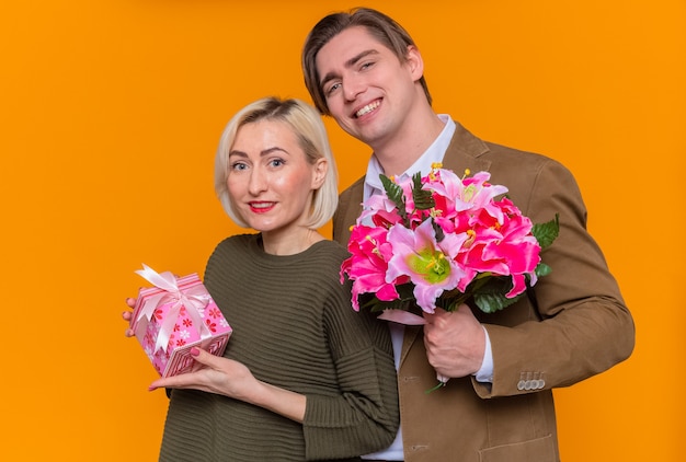 オレンジ色の壁の上に立って国際女性の日を祝う一緒に愛に幸せなプレゼントと花の花束と女性を保持している若い美しいカップル幸せな男 無料の写真