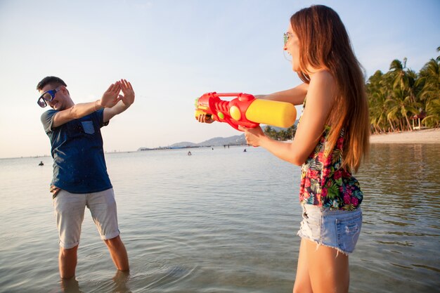 熱帯のビーチで遊ぶのが大好きな若い美しいカップル 夏休み 新婚旅行 ロマンス 日没 幸せ 楽しんで 水鉄砲 戦い 男はあきらめる ポジティブ 面白い 無料の写真