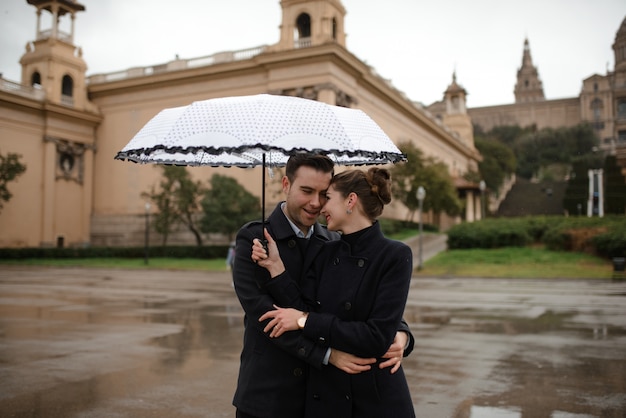 スペインのプラザで雨の中に傘の下で歩く美しい愛情のあるヒスパニックのカップル カタルーニャ国立美術館を背景にポーズをとるカップル プレミアム写真