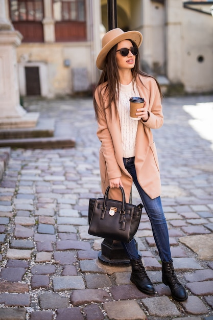 無料の写真 ハンドバッグと一杯のコーヒーが付いている通りに沿って歩く若い美しいきれいな女性