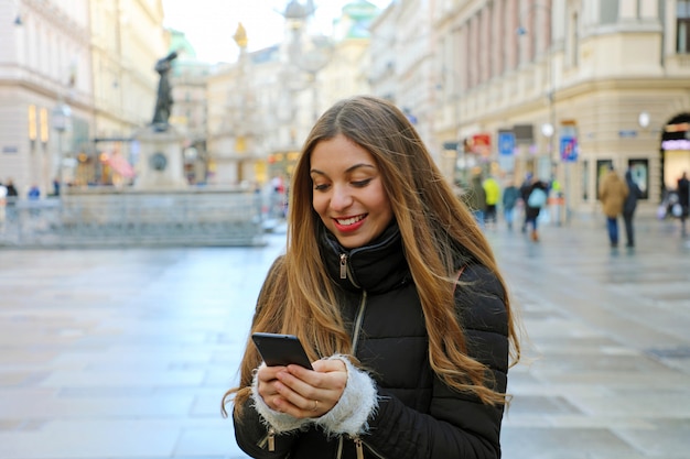 ヨーロッパの都市で屋外の電話を使用してカジュアルな冬の服を着ている若い美しい女性 オーストリアのウィーンのメインストリートで携帯電話を持つ少女 プレミアム写真
