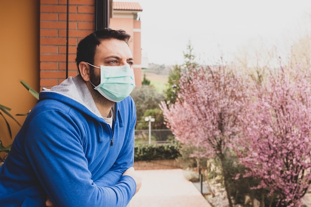Young caucasian man with mask looking out on the terrace of the house during the quarantine due to t