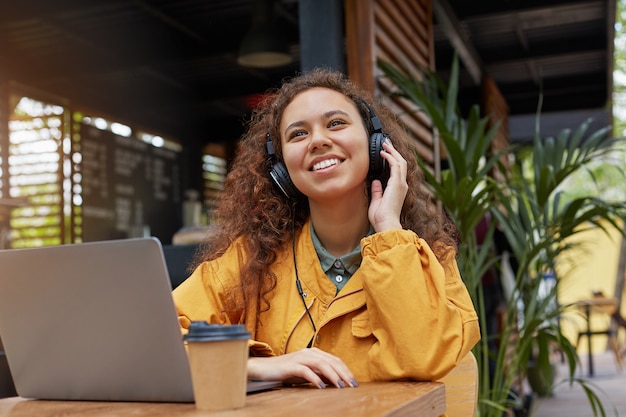 Young dark skinned curly student woman siting on a cafe terrace, listens to music and dreaming about weekend party, wearing in yellow coat, drinking coffee, works at a laptop. Free Photo