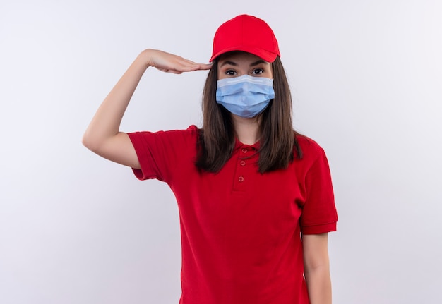 赤い帽子に赤いtシャツを着ている若い配達の女の子はフェイスマスクを着て 孤立した白い背景に敬礼を与える 無料の写真
