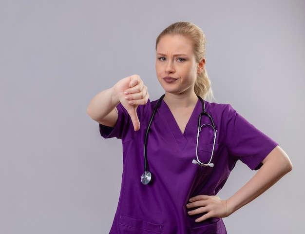 紫色の医療用ガウンと聴診器を身に着けている若い医者は 孤立した白い壁に親指を下ろします 無料の写真