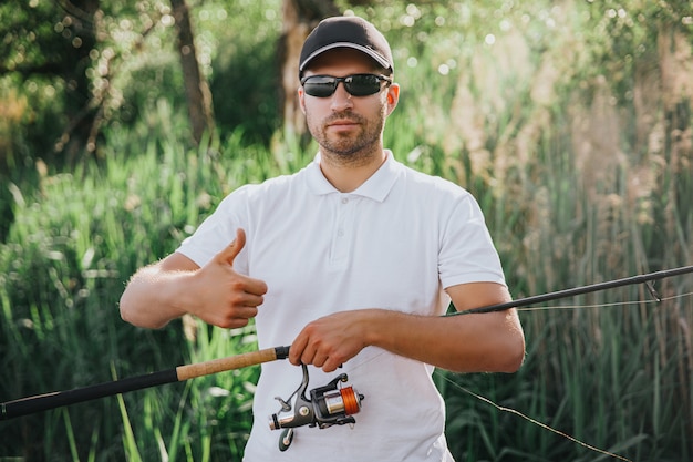 湖や川で釣りをする若い漁師 男は一人で立ち 大きな親指を立てます 下にリールを付けて長いロッドを運びます 魚や水の狩猟を捕まえる前にカメラでポーズをとる 釣り竿 プレミアム写真