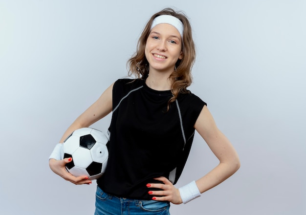 白い壁の上に立って自信を持って笑顔のサッカーボールを保持しているヘッドバンドと黒のスポーツウェアの若いフィットネスの女の子 無料の写真