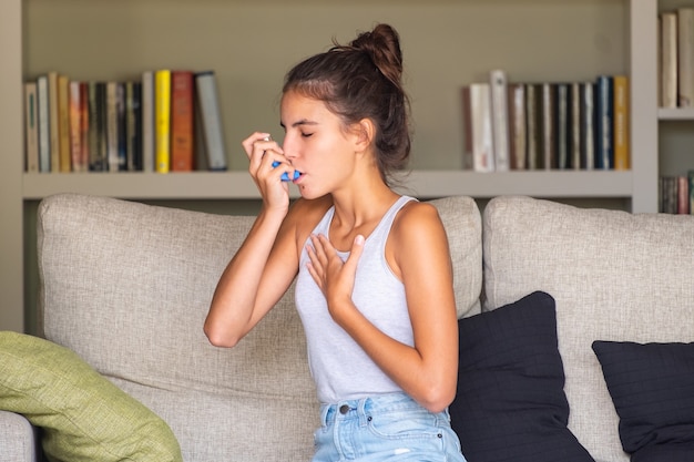 Young girl having an asthma attack and using an inhaler sitting in a sofa at home Premium Photo