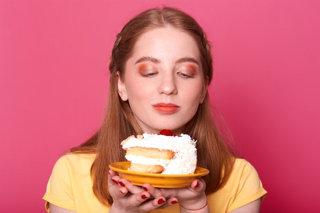 若い女の子 ピンクに分離されたバースデーケーキでプレートを見て おいしいデザートを食べたい 黄色のtシャツを着て 完璧なヘアスタイル ポーズを持っています 無料の写真