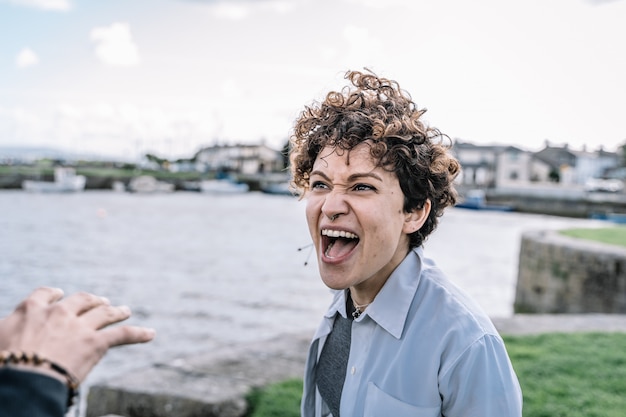 Young girl with curly hair and a piercing nose arguing with her partner shouting with expressive gestures with the sea out of focus. Free Photo