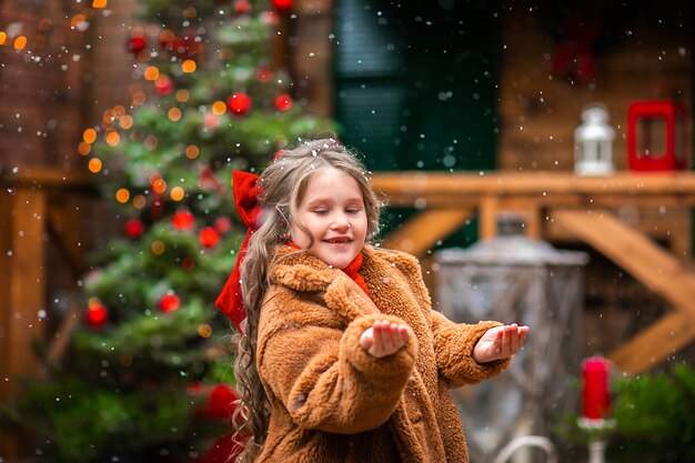 クリスマスツリーや装飾に対して雪を楽しんでいる赤いクリスマスの弓を持つ少女 プレミアム写真