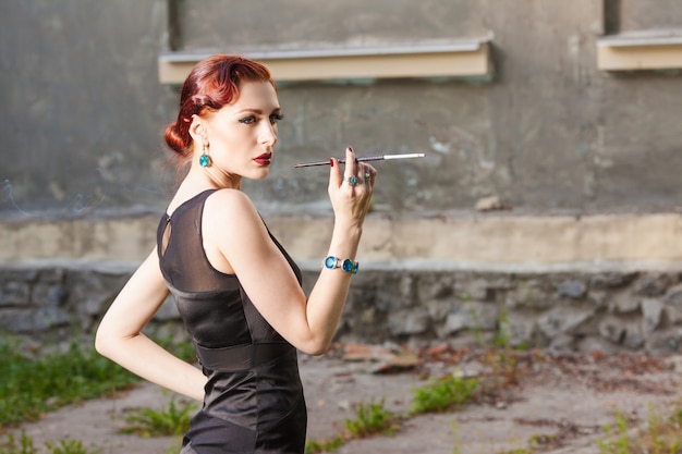 Young girl with red hair in black dress smokes cigarette in the ...