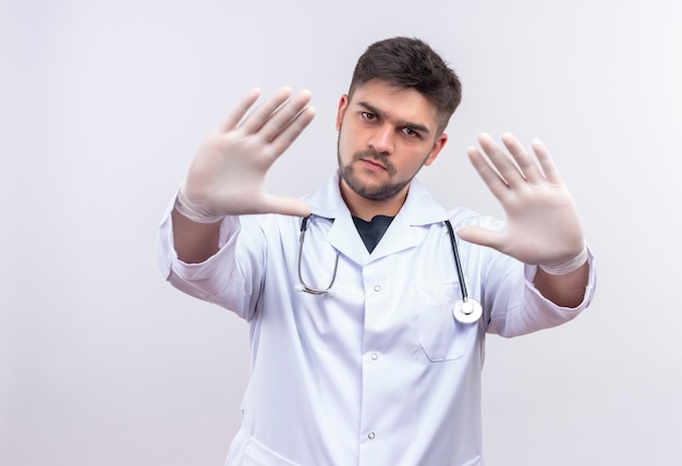 白い医療用ガウン白い医療用手袋と聴診器を身に着けている若いハンサムな医師は 白い壁の上に立っている手で一時停止の標識を怒って示しています 無料の写真