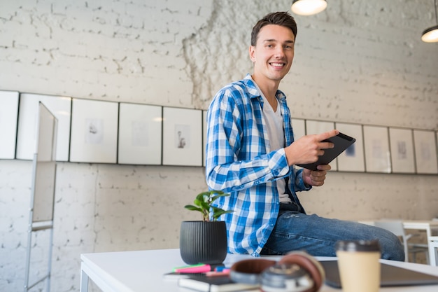 Young handsome man in chekered shirt sitting on table using tablet computer in co-working office Free Photo