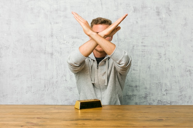 Young handsome man holding a gold ingot on a table keeping two arms crossed, denial concept. Premium Photo