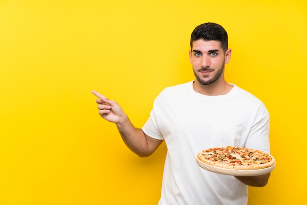 Young handsome man holding a pizza over isolated yellow wall pointing ...