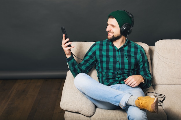Young hipster handsome bearded man sitting on a couch at home, listening to music on headphones, looking smartphone, green checkered shirt, entertainment Free Photo