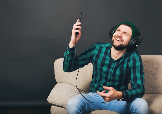Young hipster handsome bearded man sitting on a couch at home, listening to music on headphones, looking smartphone, green checkered shirt, entertainment Free Photo