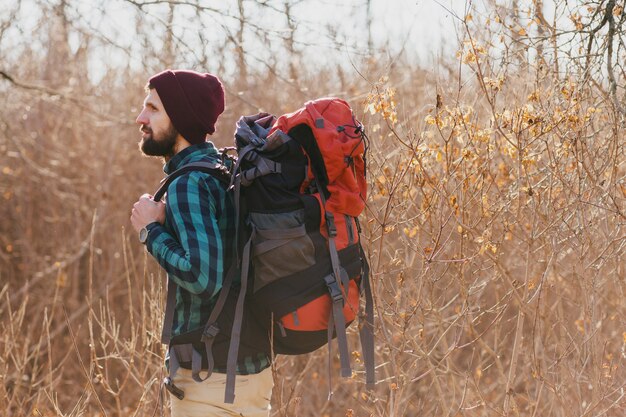 Young hipster man traveling with backpack in autumn forest wearing checkered shirt and hat, active tourist, exploring nature in cold season Free Photo