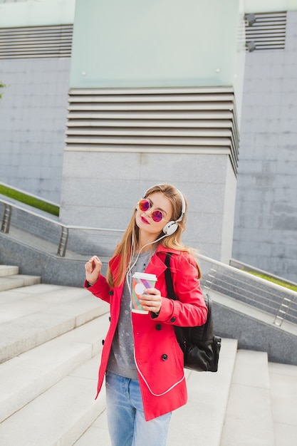 ピンクのコートを着た若いヒップスターの女性 バックパックとヘッドフォンで音楽を聴いているコーヒーと通りのジーンズ 無料の写真