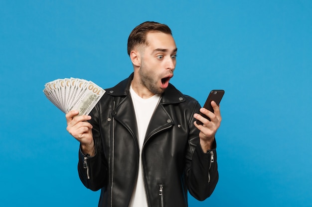 Young man in black leather jacket white t-shirt holding fan of cash money in dollar banknotes, cellphone isolated on blue wall background studio portrait. people lifestyle concept. mock up copy space. Free Photo