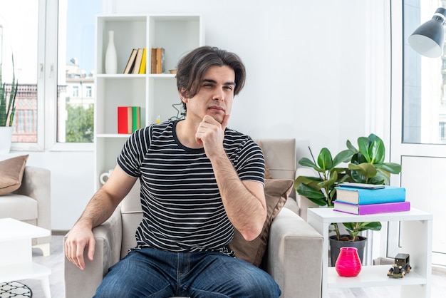 Free Photo Young Man In Casual Clothes Sitting On The Chair With Pensive Expression With Hand On Chin In Light Living Room