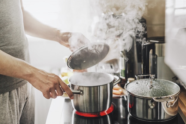 Young man cooking fresh food at home and opening lid of steaming pot. Free Photo
