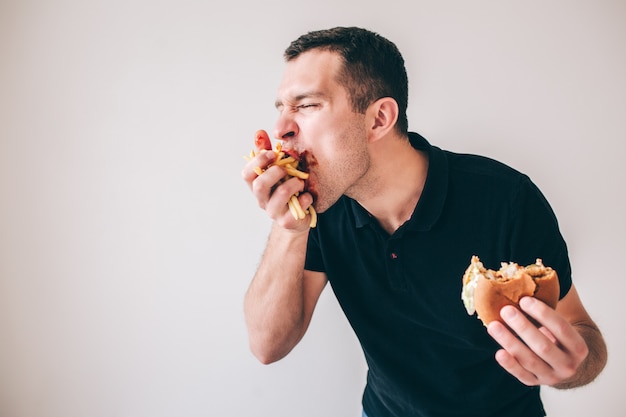 若い男が白い壁に分離されました 手に持ったフライドポテトを食べて食べる大食い人 別のかまバーガー おいしいおいしいスナックや食事 プレミアム写真