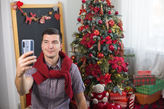 快適な服を着て自宅のクリスマスツリーの近くの若い男は スマートフォンの画面の前でこんにちは さようならのサインとして手を振る クリスマスと新年のビデオ接続おめでとうございます プレミアム写真