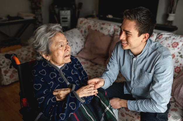 Young man sitting next to an old sick aged woman in wheelchair taking her hands while talking and smiling. family, home care concept. Premium Photo