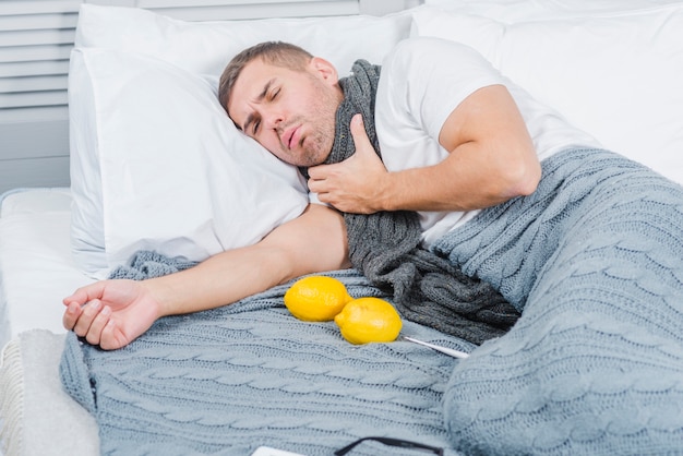 Young man suffering from sore throat lying on bed with lemon and thermometer Free Photo