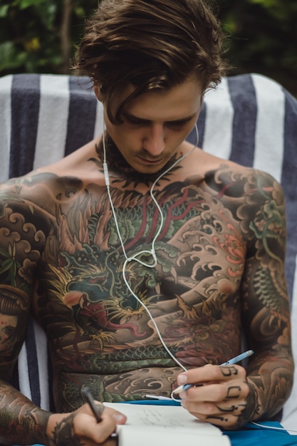 Free Photo A Young Man In Tattoos Wearing Headphones Listens To Music And Draws In A Notebook