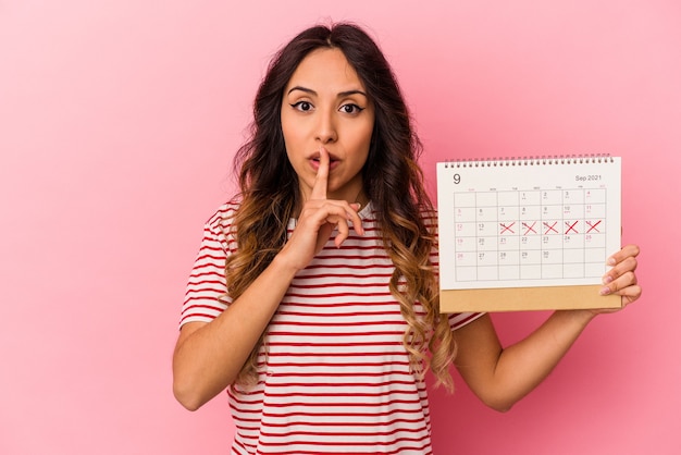 Young mexican woman holding a calendar isolated on pink keeping a secret or asking for silence. Premium Photo