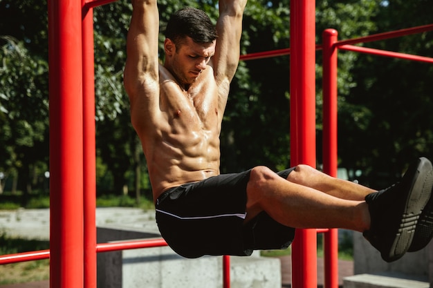 Young muscular shirtless caucasian man doing pull-ups on horizontal bar at playground in sunny summer's day. training his upper body outdoors. concept of sport, workout, healthy lifestyle, wellbeing. Free Photo