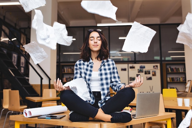 Young pretty joyful brunette woman meditating on table surround work stuff and flying papers. cheerful mood, taking a break, working, studying, relaxation, true emotions. Free Photo