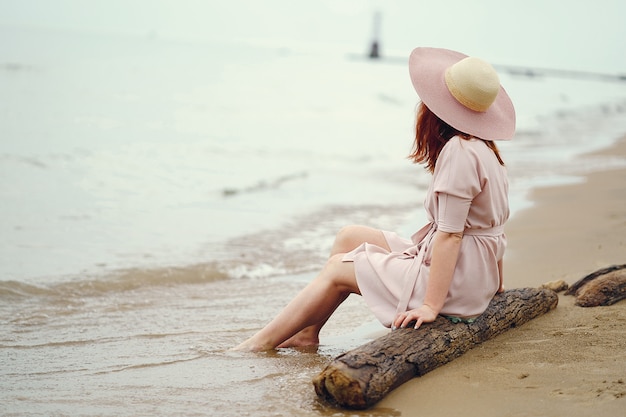A young redheaded girl in a large round hat and pink dress sitting on the beach near ocean Free Photo