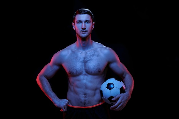 孤立して黒い背景に対してカメラの前に立っているサッカーボールを持つ若い真面目な筋肉の上半身裸のサッカー選手 プレミアム写真