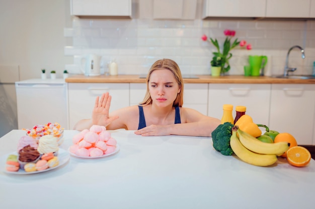 Young serious woman in blue t-shirt choosing between fresh fruit vegetables or sweets in the kitchen. choice between healthy and unhealthy food. dieting. diet. healthy food Premium Photo