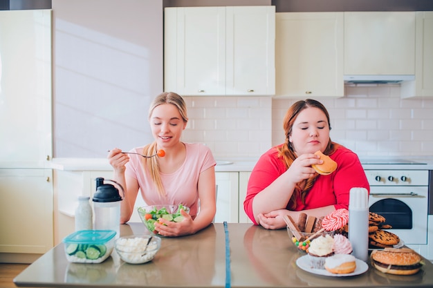  Young slim and overweight women sit at table in kitchen and eat food. healthy and unhealthy side. l