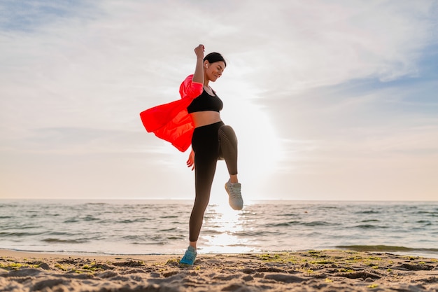 Young smiling attractive slim woman doing sports in morning sunrise jumping on sea beach in sports wear, healthy lifestyle, listening to music on earphones, wearing pink windbreaker jacket, having fun Free Photo