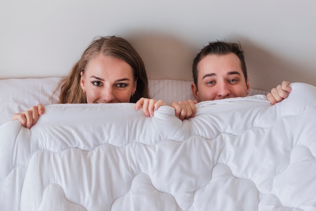 Young Smiling Couple Lying On Bed And Peeping Out From Duvet